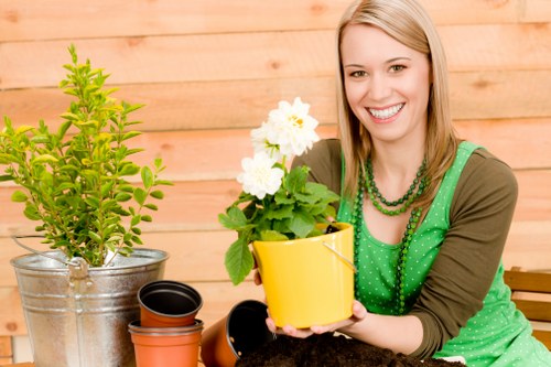 A woman with light brown hair and a bright smile is sitting in an outdoor garden space with a wooden fence in the background. She is holding a yellow flower pot containing a healthy, blooming white flowering plant, possibly a geranium or similar. In front of her on the table are various gardening items, including empty plant pots of black and terracotta colours, and a small potted shrub with vibrant green leaves in a metallic container. The garden area features a neatly maintained lawn with dense, green grass occupying the foreground. The scene is well-lit, indicating a clear day with natural sunlight. The overall setting suggests a tidy, well-kept outdoor space suitable for gardening activities, with elements that align with professional lawn care and gardening services in Camden Town, such as Lawn Mowing Camden Town, serving residential gardens and landscaped yards in the local postcode area.